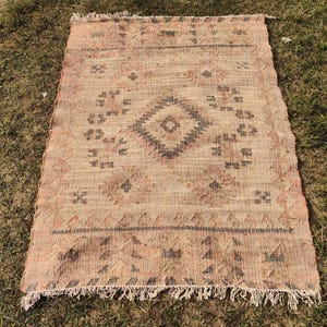 Puede incluir: Alfombra rectangular beige con un patr&oacute;n de diamante geom&eacute;trico en el centro. La alfombra tiene flecos en el borde inferior y est&aacute; colocada sobre una superficie de hierba. El dise&ntilde;o incluye detalles en negro y marr&oacute;n.