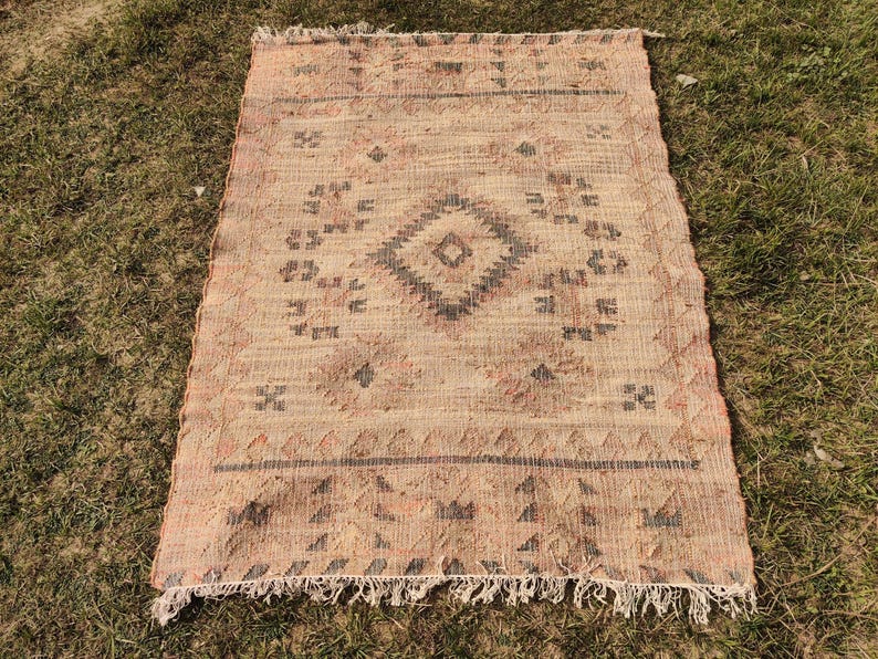 Puede incluir: Alfombra rectangular beige con un patr&oacute;n de diamante geom&eacute;trico en el centro. La alfombra tiene flecos en el borde inferior y est&aacute; colocada sobre una superficie de hierba. El dise&ntilde;o incluye detalles en negro y marr&oacute;n.