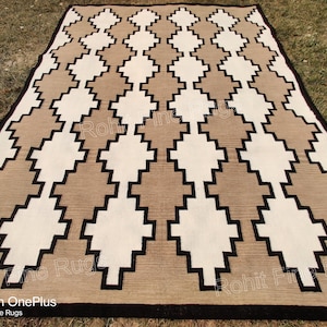 Pode incluir: Um tapete geométrico castanho e branco com detalhes pretos. O tapete tem um padrão de diamante com um centro branco e bordas castanhas. O tapete é feito de lã e é tecido à mão.
