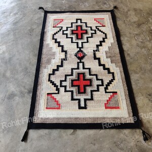 May include: Rectangular rug with a geometric pattern in beige, black, and red. The rug features a central design with two red crosses and a black border with tassels at the corners. The rug is placed on a concrete surface.