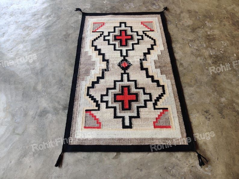 May include: Rectangular rug with a geometric pattern in beige, black, and red. The rug features a central design with two red crosses and a black border with tassels at the corners. The rug is placed on a concrete surface.