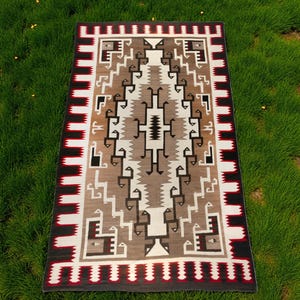 Può includere: Tappeto rettangolare con un motivo geometrico nei toni del marrone, bianco, nero e grigio. Il tappeto ha un bordo con strisce rosse e bianche alternate. Il design presenta forme e linee stilizzate, creando un motivo simmetrico e dettagliato.