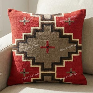 May include: Red decorative pillow with a geometric pattern in brown, gray, and cream. The pillow has a textured appearance and is placed on a light-colored sofa. The pillow's design features a central cross motif.