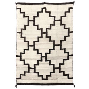 Op de afbeelding: Een wit Navajo-kleed met een geometrisch patroon van zwarte vierkanten en rechthoeken. Het patroon is een herhalend ontwerp van in elkaar grijpende vierkanten en rechthoeken.
