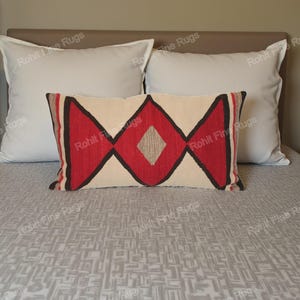 Op de afbeelding: Een decoratief lendenkussen met een geometrisch ontwerp in rood, zwart en beige. Het kussen heeft een centrale ruitvorm. Twee witte vierkante kussens flankeren het kussen op een bed met een grijze sprei met patroon.