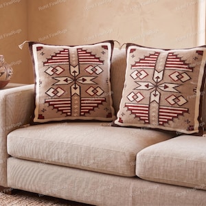May include: Two square throw pillows featuring a tan base with a geometric pattern in dark brown, white, and deep red. Each pillow has a dark brown border and small tassels at the corners, displaying a symmetrical design of stepped lines and diamonds.