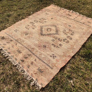 Puede incluir: Una alfombra rectangular tejida a mano con un aspecto descolorido y vintage. La alfombra presenta un patr&oacute;n de diamante central y dise&ntilde;os geom&eacute;tricos en tonos beige, marr&oacute;n y rojo. Los bordes tienen un detalle de flecos y la alfombra est&aacute; colocada sobre una superficie de hierba.