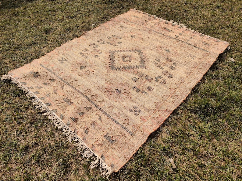 Puede incluir: Una alfombra rectangular tejida a mano con un aspecto descolorido y vintage. La alfombra presenta un patr&oacute;n de diamante central y dise&ntilde;os geom&eacute;tricos en tonos beige, marr&oacute;n y rojo. Los bordes tienen un detalle de flecos y la alfombra est&aacute; colocada sobre una superficie de hierba.