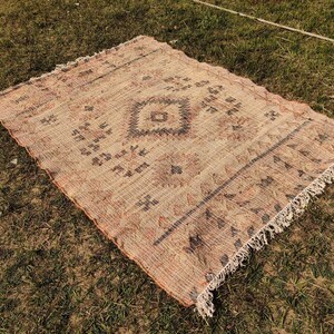Puede incluir: Una alfombra rectangular tejida con un patr&oacute;n de diamante central y bordes decorativos. La alfombra presenta una paleta de colores beige y marr&oacute;n con flecos en los bordes. La alfombra est&aacute; colocada sobre hierba verde.