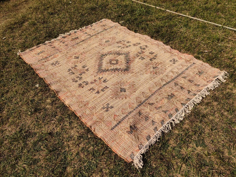 Puede incluir: Una alfombra rectangular tejida con un patr&oacute;n de diamante central y bordes decorativos. La alfombra presenta una paleta de colores beige y marr&oacute;n con flecos en los bordes. La alfombra est&aacute; colocada sobre hierba verde.