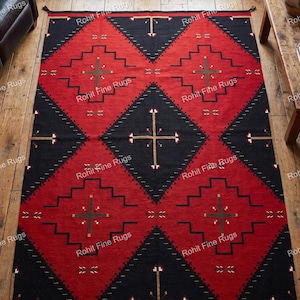 Op de afbeelding: Een rechthoekig rood en zwart patroonkleed met geometrische ontwerpen. Het kleed heeft een herhalend ruitpatroon met getrapte randen en kruisachtige motieven. Het kleed ligt op een houten vloer.