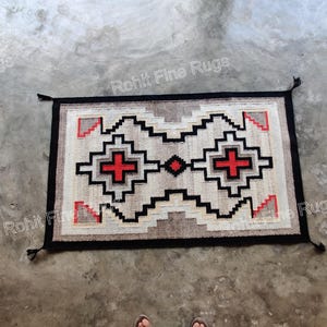 May include: Rectangular rug with a geometric pattern in beige, black, and red. The rug features a central design with cross-like shapes and a black border with tassels. The rug is placed on a concrete surface.
