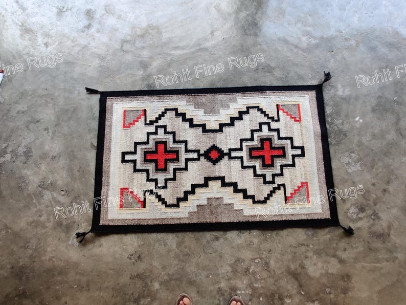 May include: Rectangular rug with a geometric pattern in beige, black, and red. The rug features a central design with cross-like shapes and a black border with tassels. The rug is placed on a concrete surface.