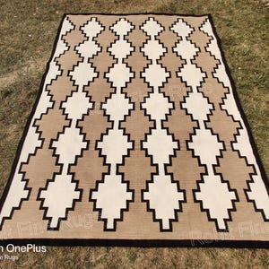 Könnte beinhalten: Ein brauner und weißer Teppich mit Rautenmuster und schwarzen Rändern. Der Teppich hat ein geometrisches Design und ist aus gewebten Fasern gefertigt.