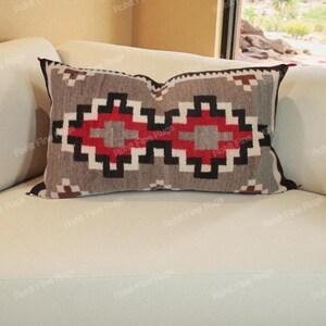 Pode incluir: Almofada decorativa retangular com um padrão geométrico em tons de cinza, vermelho e preto. A almofada está colocada em um sofá branco, mostrando uma estética de design do sudoeste.