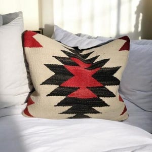 Puede incluir: Una almohada blanca con un patrón geométrico rojo, negro y blanco. La almohada está sobre una cama blanca con sábanas blancas.