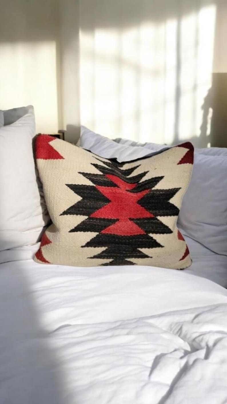 May include: A white pillow with a red, black, and white geometric pattern. The pillow is on a white bed with white sheets.