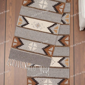 May include: A rectangular gray and cream-colored runner rug with geometric patterns and fringe. The rug features brown and black zigzag and cross designs. The rug is displayed on a wooden floor.