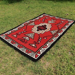Op de afbeelding: Een rood, zwart en wit patroonteppich met een geometrisch ontwerp. Het tapijt ligt op een groene grasmat.