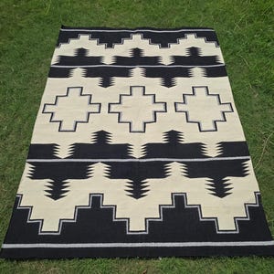 Puede incluir: Alfombra rectangular con un patrón geométrico en negro y crema. La alfombra presenta formas y líneas repetidas, creando un diseño simétrico. La alfombra está colocada sobre una superficie de hierba verde.