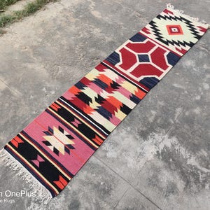 Puede incluir: Una alfombra larga y estrecha con un patrón geométrico en tonos rojos, blancos, negros y azules. La alfombra tiene flecos en ambos extremos. "Shot on OnePlus By Rohit Fine Rugs" está impreso en la parte inferior de la imagen.
