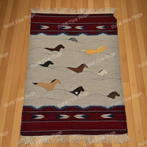 Puede incluir: Una alfombra rectangular con un fondo gris que presenta un patrón de caballos corriendo en varios colores. La alfombra tiene un borde rojo con diseños geométricos y flecos blancos.