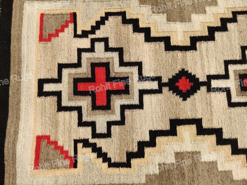 May include: Close-up of a woven rug with a geometric pattern. The design features black, beige, and red shapes, including crosses and zigzags. The rug has a textured appearance and a border of black and beige stripes.