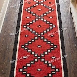 Puede incluir: Una alfombra rectangular roja con borde negro y un patrón geométrico de diamantes blancos y negros. La alfombra está colocada sobre un suelo de madera oscura. Se ven borlas en los extremos de la alfombra.
