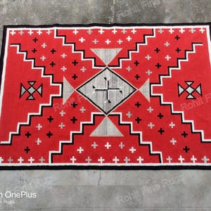 Op de afbeelding: Een rood, wit en zwart Navajo-kleed met een geometrisch patroon. Het kleed heeft een centrale diamantvorm met een kruis erin. De diamant wordt omringd door een reeks concentrische vierkanten en driehoeken.