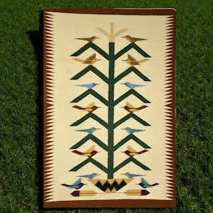 Puede incluir: Alfombra rectangular tejida con fondo crema y diseño central de árbol. El árbol es verde con pájaros marrones y azules posados en sus ramas. El borde es marrón con un patrón en zigzag.