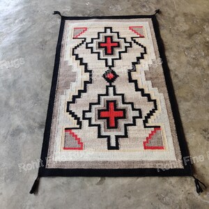 May include: Rectangular rug with a geometric pattern in beige, black, red, and gray. The rug features a central design with red crosses and zigzag patterns. The border is black, and the rug has tassels at the corners.