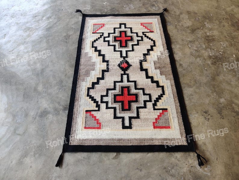 May include: Rectangular rug with a geometric pattern in beige, black, red, and gray. The rug features a central design with red crosses and zigzag patterns. The border is black, and the rug has tassels at the corners.