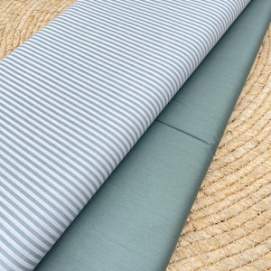 Könnte beinhalten: Zwei Stoffmuster. Eines hat ein hellblau-weißes Streifenmuster, das andere ist einfarbig hellblau. Die Stoffe sind gefaltet und geschichtet, wodurch ihre Texturen und Farben hervorgehoben werden. Der Hintergrund ist eine gewebte, natürlich getönte Matte.