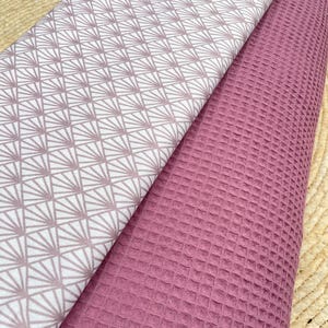 Könnte beinhalten: Zwei Stoffmuster, übereinandergelegt. Eines zeigt ein weißes Muster mit einem geometrischen Fächerdesign in Mauve, das andere eine einfarbige Waffelstruktur in Mauve. Die Stoffe sind gefaltet und zeigen ihre unterschiedlichen Texturen und Farben.