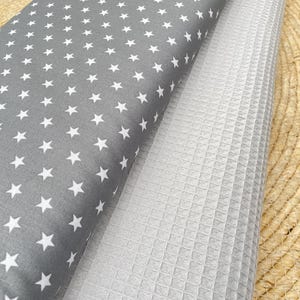 Puede incluir: Dos rollos de tela. Uno es gris con patrones de estrellas blancas. El otro es blanco con una textura de tejido de gofre. Las telas están enrolladas y colocadas sobre una superficie tejida.