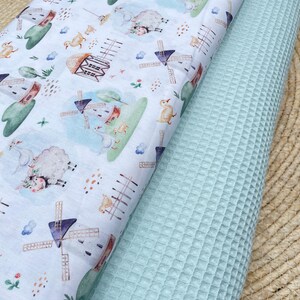 Puede incluir: Primer plano de dos rollos de tela. Uno presenta un fondo blanco con un estampado caprichoso de molinos de viento, ovejas y patos en colores pastel. El otro es una tela de gofre azul claro liso.