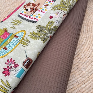 Könnte beinhalten: Zwei Stoffrollen. Eine hat einen hellgrünen Hintergrund mit einem sich wiederholenden Muster aus Zeichentricktieren, Pflanzen und einem Radio. Die andere ist ein einfarbiger brauner Waffelstoff. Die Stoffe liegen auf einer gewebten Matte.