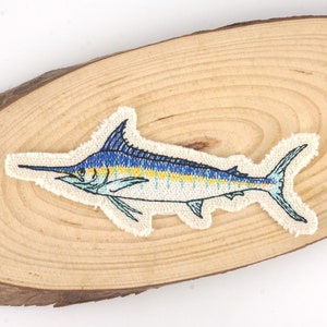 以下が含まれることがあります： 薄い茶色の木製の表面に、青いカジキの刺繍パッチが施されています