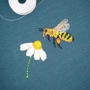 May include: Embroidered bee and daisy on teal fabric. The bee is yellow and black with detailed wings, appearing to hover near the daisy. The daisy has white petals and a yellow centre, with a green stem. White thread is visible.