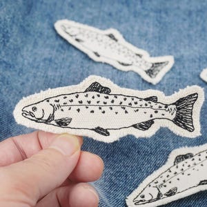 以下が含まれることがあります： 青いデニムの背景に、刺繍されたアイロン接着パッチが4つ。サーモンは黒い糸で縁取られ、黒い斑点がある。