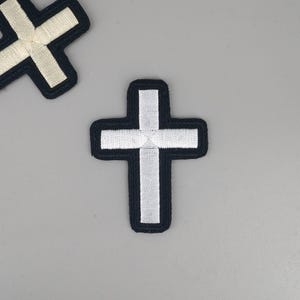 May include: Two embroidered cross patches. The crosses have a white center with a black border. One cross is in the shape of a plus sign, and the other is an X shape. The patches are on a gray background.