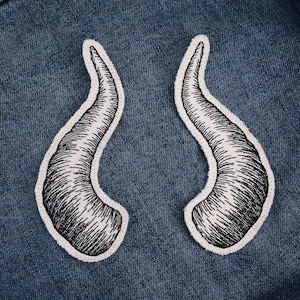 Może przedstawiać: Dwie haftowane naszywki w kształcie zakrzywionych rogów. Rogi są czarne z białymi przeszyciami i białym obramowaniem. Znajdują się na niebieskim tle z dżinsu.