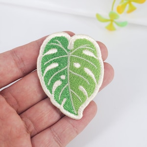 May include: A green and white embroidered patch of a monstera leaf. The patch is about the size of a hand.