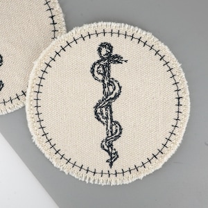 May include: Two circular, off-white fabric coasters. Each has a black embroidered caduceus symbol, a staff with two snakes. The edges are finished with a black stitched border, giving a handmade look. The background is a light grey.