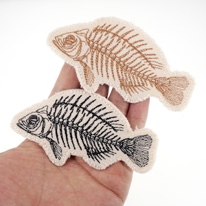 May include: Two embroidered fish skeleton patches. One is brown, the other is black, both on a cream-coloured background. The patches are detailed with stitched lines, depicting the bones and shape of a fish. The patches are held in a hand.