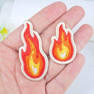 May include: Two embroidered iron-on patches in the shape of flames. The larger patch is red, orange, and yellow with a white outline. The smaller patch is the same colours but smaller in size.