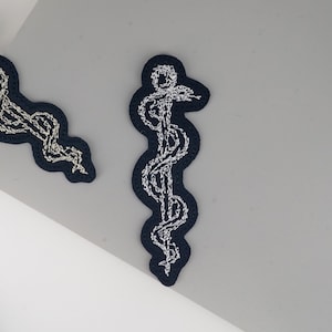 Op de afbeelding: Twee marineblauwe geborduurde patches met een wit caduceus-symbool. De patches hebben een golvende, organische vorm. De witte borduursels vallen op tegen de donkerblauwe achtergrond. De patches zijn waarschijnlijk voor medisch personeel.