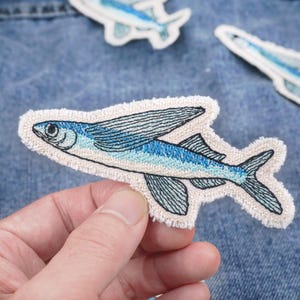 May include: Embroidered patches featuring a flying fish design. The patches are primarily blue and white, with detailed stitching and a cream-colored border. The patches are shown on a denim background.