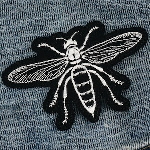 Puede incluir: Parche bordado de abeja en blanco y negro. El parche presenta un diseño detallado de abeja con costuras blancas sobre un fondo negro. Probablemente está hecho de tela y está diseñado para coser o planchar en ropa o accesorios.