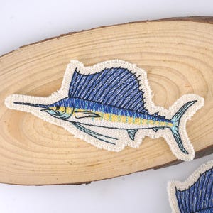 Puede incluir: Un parche bordado de un pez marlin azul y amarillo sobre una superficie de madera marrón claro.